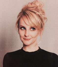 a woman with blonde hair wearing a black shirt and looking at the camera while standing in front of a white wall