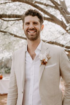 a man in a white shirt and tan jacket smiles at the camera while wearing a flower boutonniere