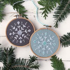 two cross stitch snowflakes hanging from evergreen needles on a white wooden background