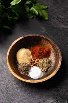 spices in a wooden bowl on a black surface next to green leaves and parsley