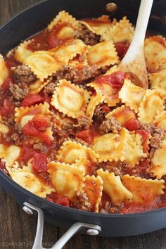 a skillet filled with pasta and meat on top of a wooden table next to a spoon