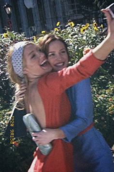 two women taking a selfie in front of some bushes and flowers with their cell phone