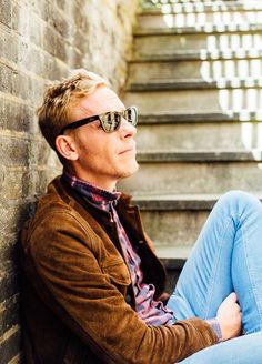 a man sitting on the ground with his legs crossed and wearing sunglasses, leaning against a brick wall