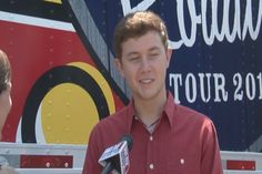 a young man standing in front of a sign talking to an interviewer about the tour