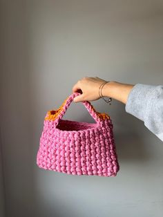 a hand holding a pink crocheted basket hanging from the wall in front of a white wall