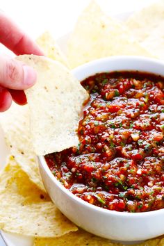 a hand dipping salsa into a bowl with tortilla chips on the side,