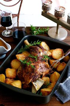 a roasting pan filled with meat and potatoes on a wooden table next to wine glasses