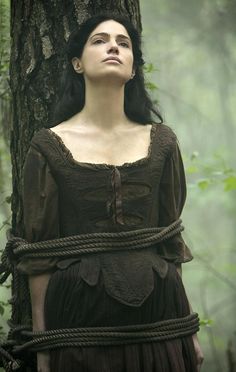 a woman standing next to a tree wearing a dress and tied up with rope around her neck