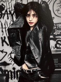 a woman with black hair wearing a leather jacket and posing in front of graffiti covered wall