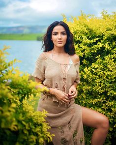 a woman posing for a photo in front of some bushes and trees with her legs crossed