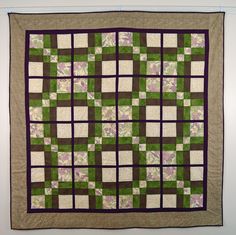 a green and brown quilt hanging on the wall next to a white wall with purple flowers