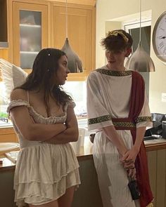 two women dressed in roman costumes standing next to each other near a kitchen counter with a clock on the wall