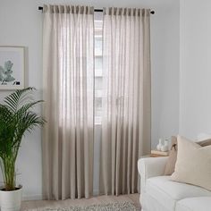 a living room with a white couch and large window covered in beige drapes next to a potted plant