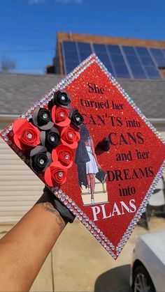 a hand holding a graduation cap decorated with red and black flowers in front of a house