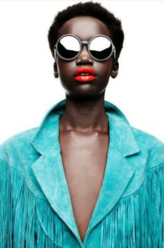 a woman with red lipstick and sunglasses on her face, wearing a blue shirt and fringed jacket