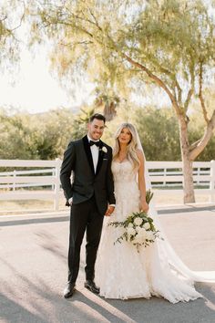 a bride and groom posing for a photo in front of a white fence with trees