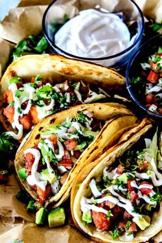 three tacos with toppings on top of them sitting in a paper tray next to a bowl of ranch dressing