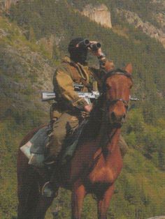 a man riding on the back of a brown horse next to a lush green hillside