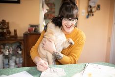 a woman holding a pug in her arms while sitting at a table with pictures on it