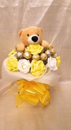 a teddy bear sitting on top of a bouquet of yellow and white roses with chocolates