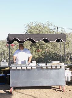 a man standing behind a table with food on it in front of some trees and bushes