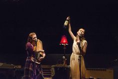 two women singing into microphones while standing on stage with other people in the background