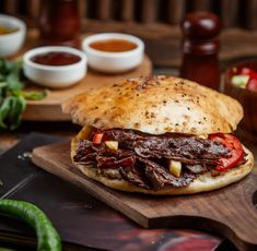 a roast beef sandwich with tomatoes and peppers on a cutting board next to dipping sauces