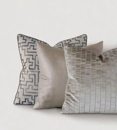 three decorative pillows in various shades of grey and gold on a white background, one with an intricate design