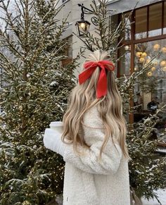 a woman standing in front of a christmas tree wearing a white coat and red bow