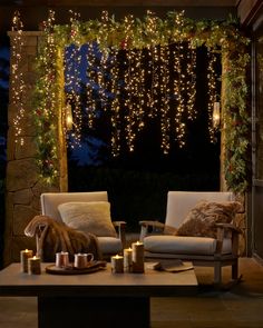 an outdoor living room with lights strung from the ceiling and two chairs in front of it