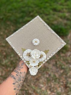 a person's hand holding up a silver and white graduation cap with flowers on it