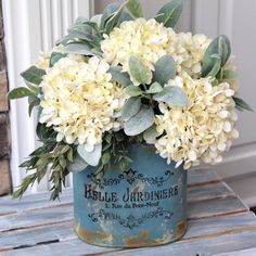 a blue vase with white flowers and greenery in it sitting on a wooden table