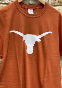 Show off your team pride in this Texas Longhorns Burnt Orange Primary Logo Short Sleeve T Shirt! This Texas Short Sleeve Tee features a screen print Longhorn logo on front chest. Make sure everyone knows you root for the Longhorns with this Burnt Orange Texas T Shirt. Hook Em Horns! Ribbed Crew Neck, Soft Cotton Solid Jersey, Finished hem and arm holes, Screen printed team graphic on center chest, Comfortable material, Fit: True to Size, 100% COTTON, Machine Washable, 8 Casual Crew Neck T-shirt For Football Season, Collegiate Fan Merchandise T-shirt For Fall, Collegiate Fall Fan Merchandise T-shirt, Brown Crew Neck Top For College, Fall Team Spirit T-shirt With Logo Print, Orange Crew Neck T-shirt With Team Name, Collegiate Crew Neck T-shirt For Fall, Orange Tops For Fan Gear With Team Spirit, Fan Gear Graphic Tee With Crew Neck