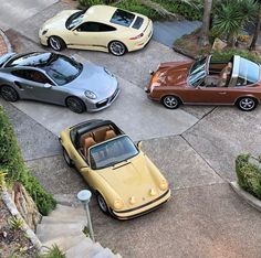 four different colored sports cars parked in a parking lot next to trees and bushes on the side walk