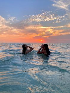 two women in the ocean at sunset with their backs to each other, one laying on her head