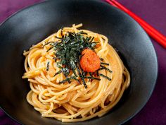 a black bowl filled with pasta and topped with an orange garnish next to chopsticks