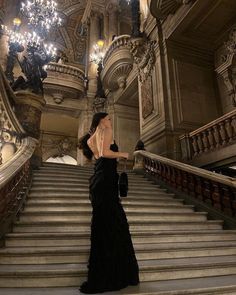 a woman in a long black dress is standing on the stairs with chandelier