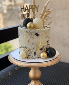 a white and gold birthday cake with black and gold decorations on a table in front of a window