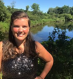 a woman standing in front of a river with trees and bushes around her, smiling for the camera