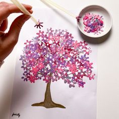 someone is painting a tree with pink, purple and blue flowers on paper next to a white bowl