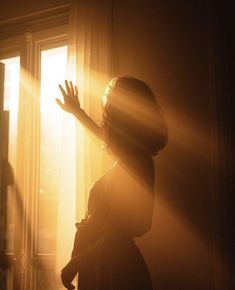 a woman standing in front of a window with her hand up to the light coming through