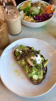 a white plate topped with food next to a bowl of salad and a cup of drink