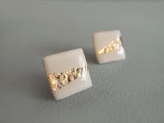 two white and gold square shaped earrings sitting on top of a gray surface with one earring in the shape of a rectangle