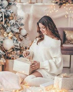 a woman sitting in front of a christmas tree holding a box with presents on it