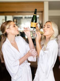 two women in white robes are drinking wine from champagne glasses while standing next to each other