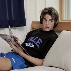 a boy laying in bed with headphones on reading a book