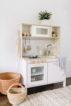 a white play kitchen with an oven, microwave and basket on the floor next to it