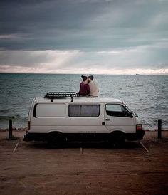 two people sitting on the roof of a van by the ocean under a cloudy sky