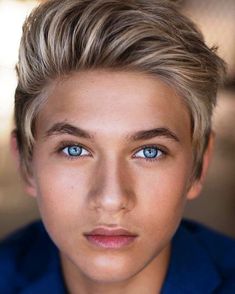 a young man with blonde hair and blue eyes looks at the camera while wearing a blue shirt