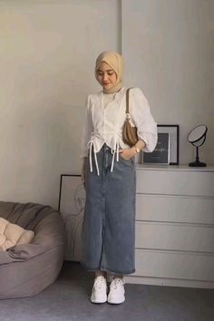 a woman standing next to a bed wearing a white shirt and grey skirt with laces